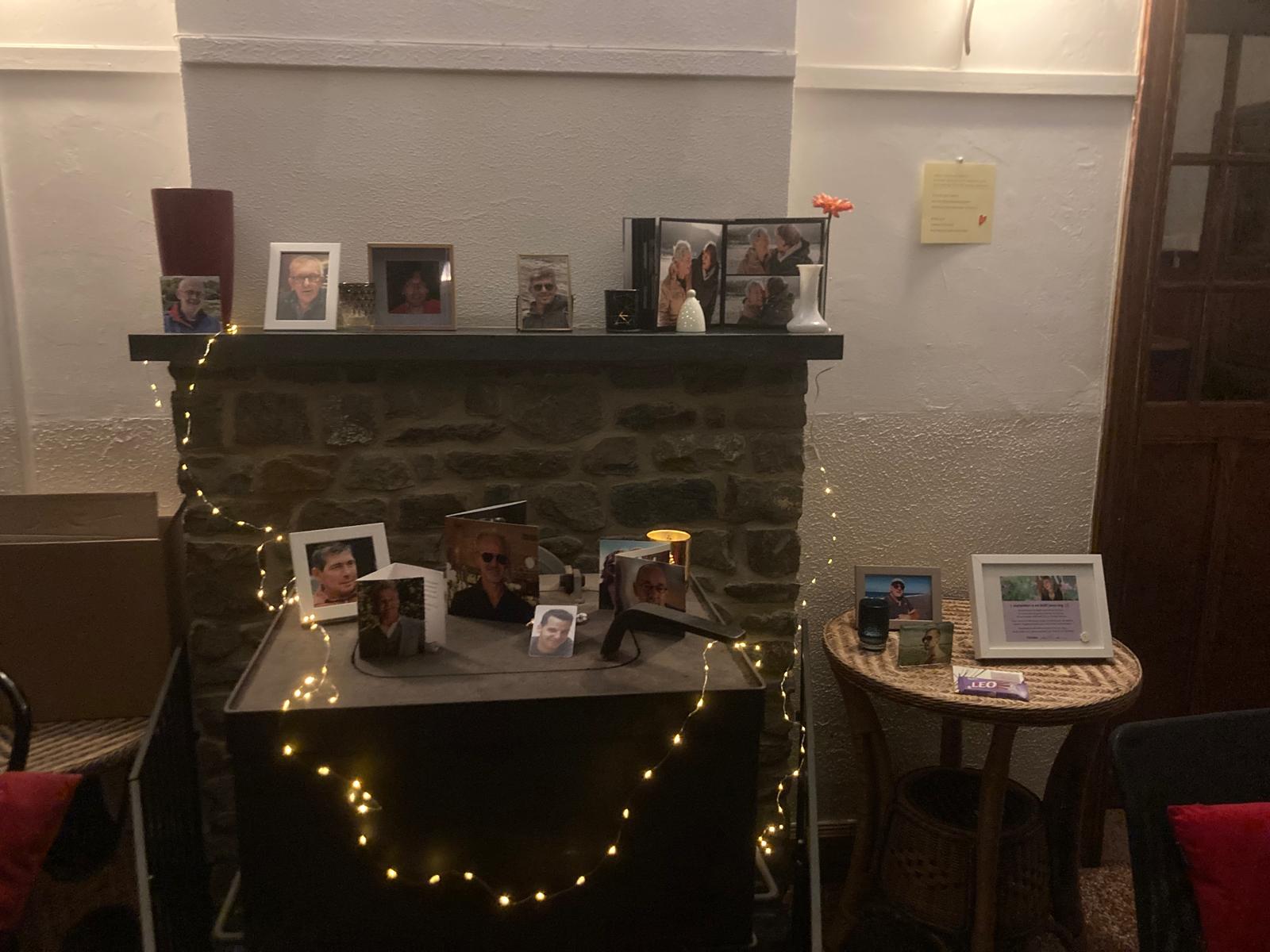 Sfeervolle herdenkingshoek met ingelijste foto’s, kaarsen en lichtsnoer op een stenen schouw.