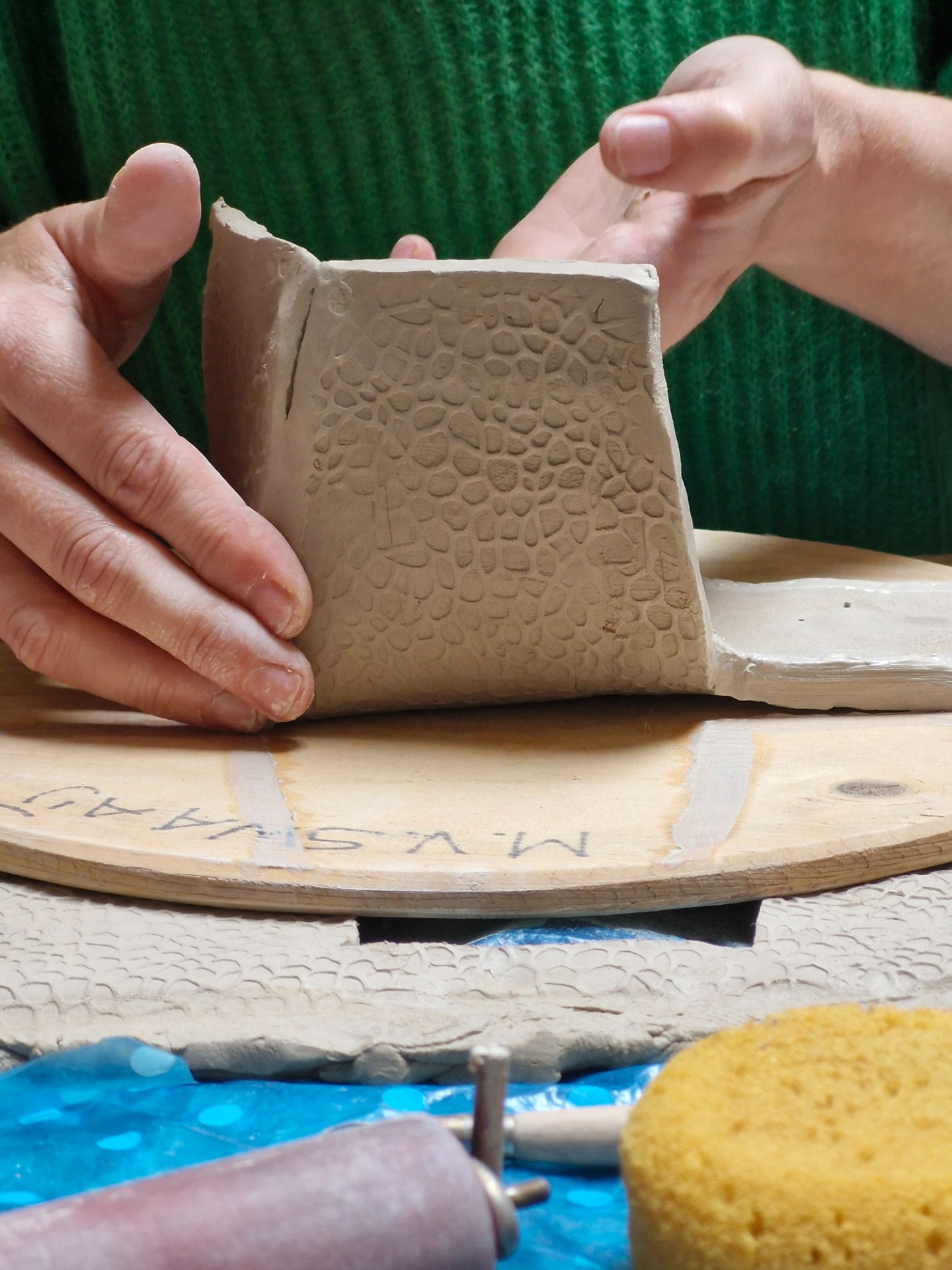 Handen houden een kleiblok met reliëf vast, terwijl het op een houten plank wordt bewerkt.