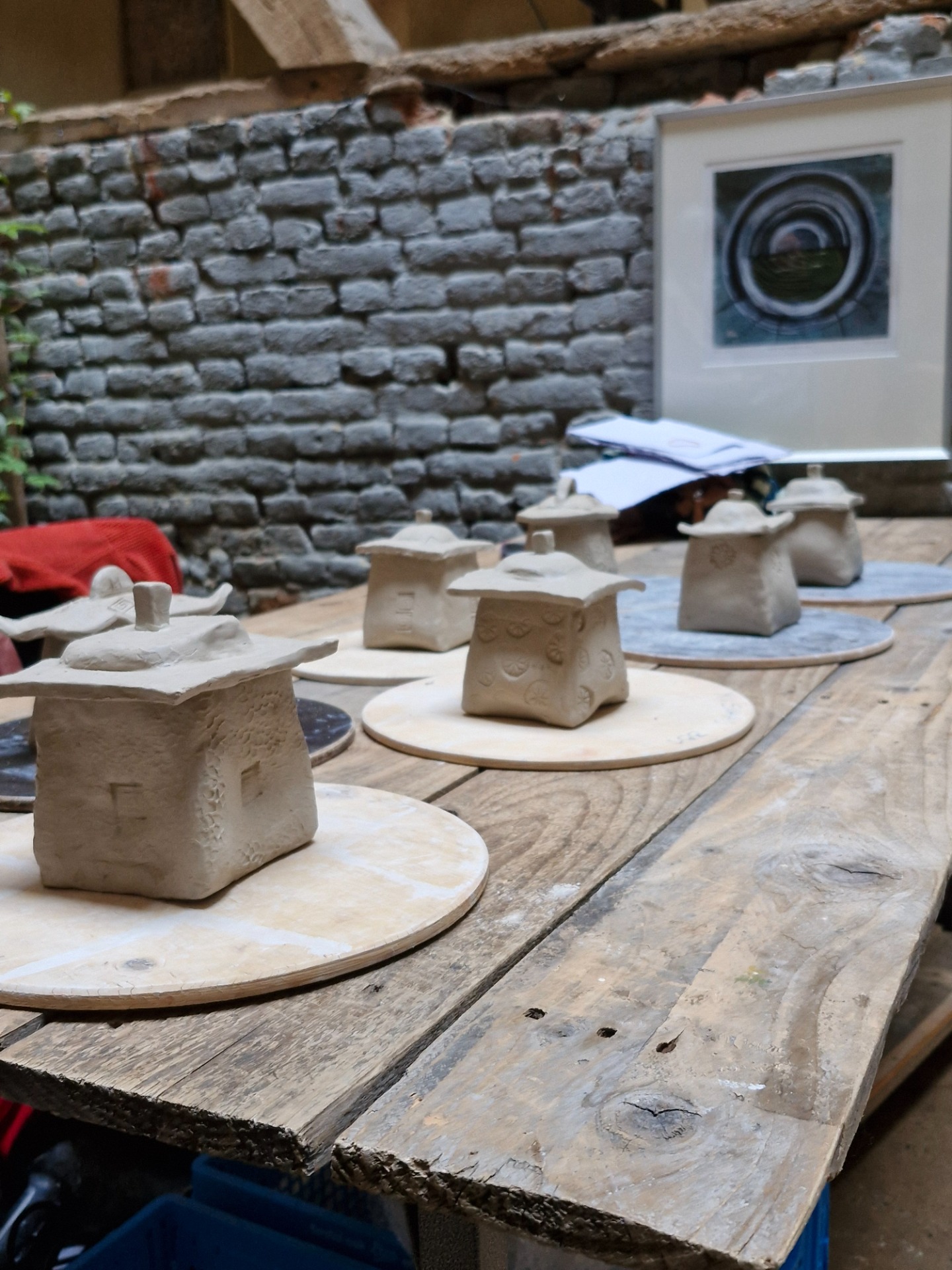 Rij van klei- of gipsvormen op ronde planken, met stenen muur en abstract kunstwerk op achtergrond.