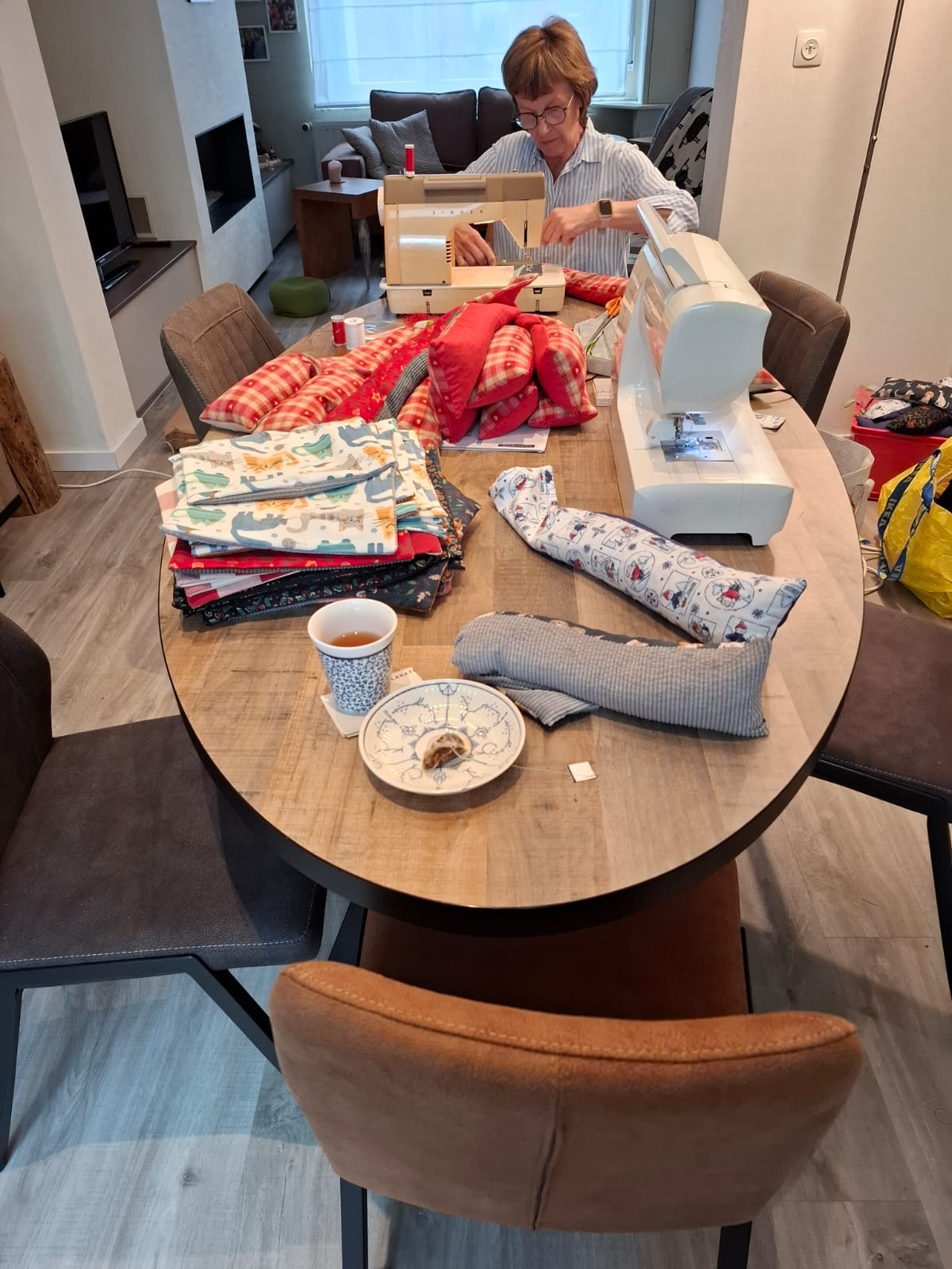 Vrouw naait aan een naaimachine aan tafel met stoffen, kussens en een kopje thee in woonkamer.