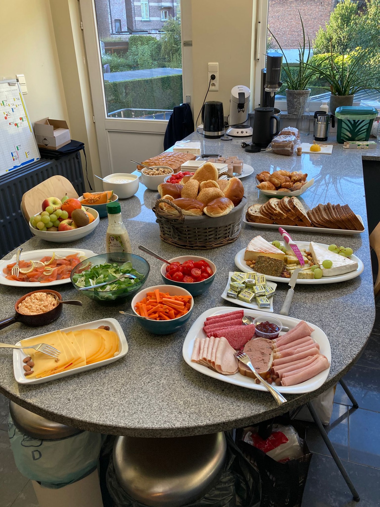 Ontbijtbuffet met borden vol beleg, broodmand, fruit op een grijze keukentafel.