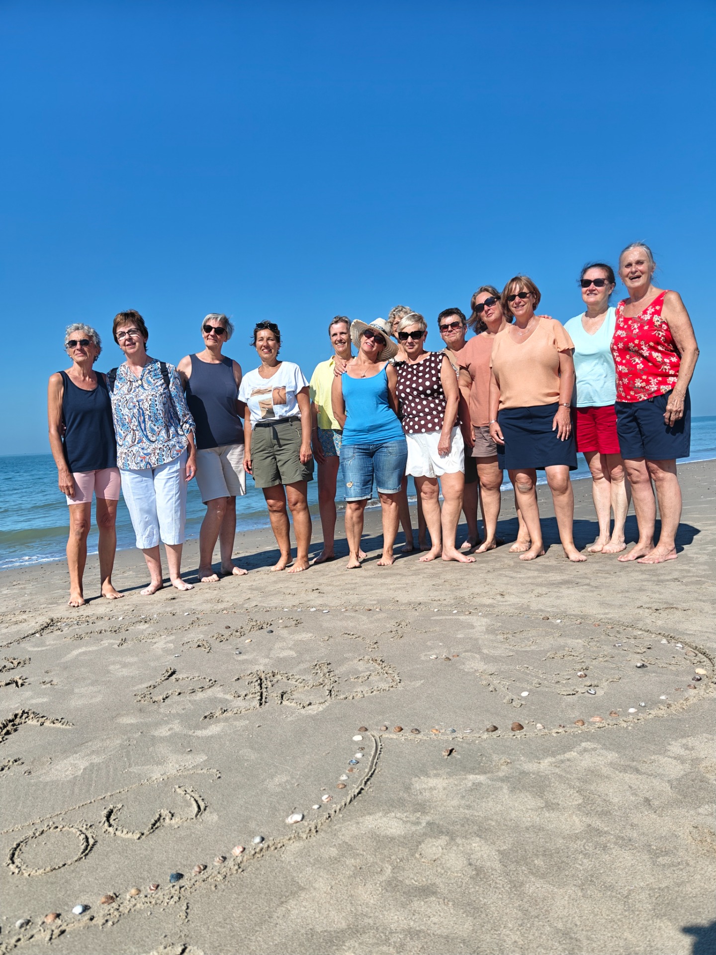 Groep poseert op een zonnig strand met blote voeten, blauwe lucht en zee op de achtergrond.