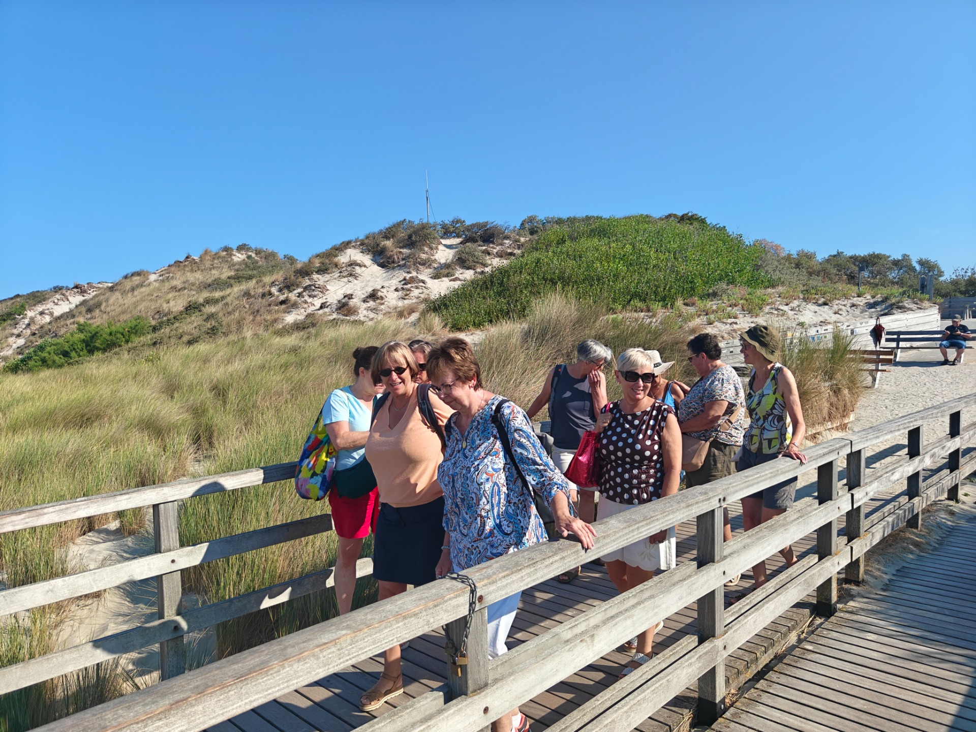 Groep poseert op houten duinpad bij strand, met duinen en helmgras onder blauwe lucht.