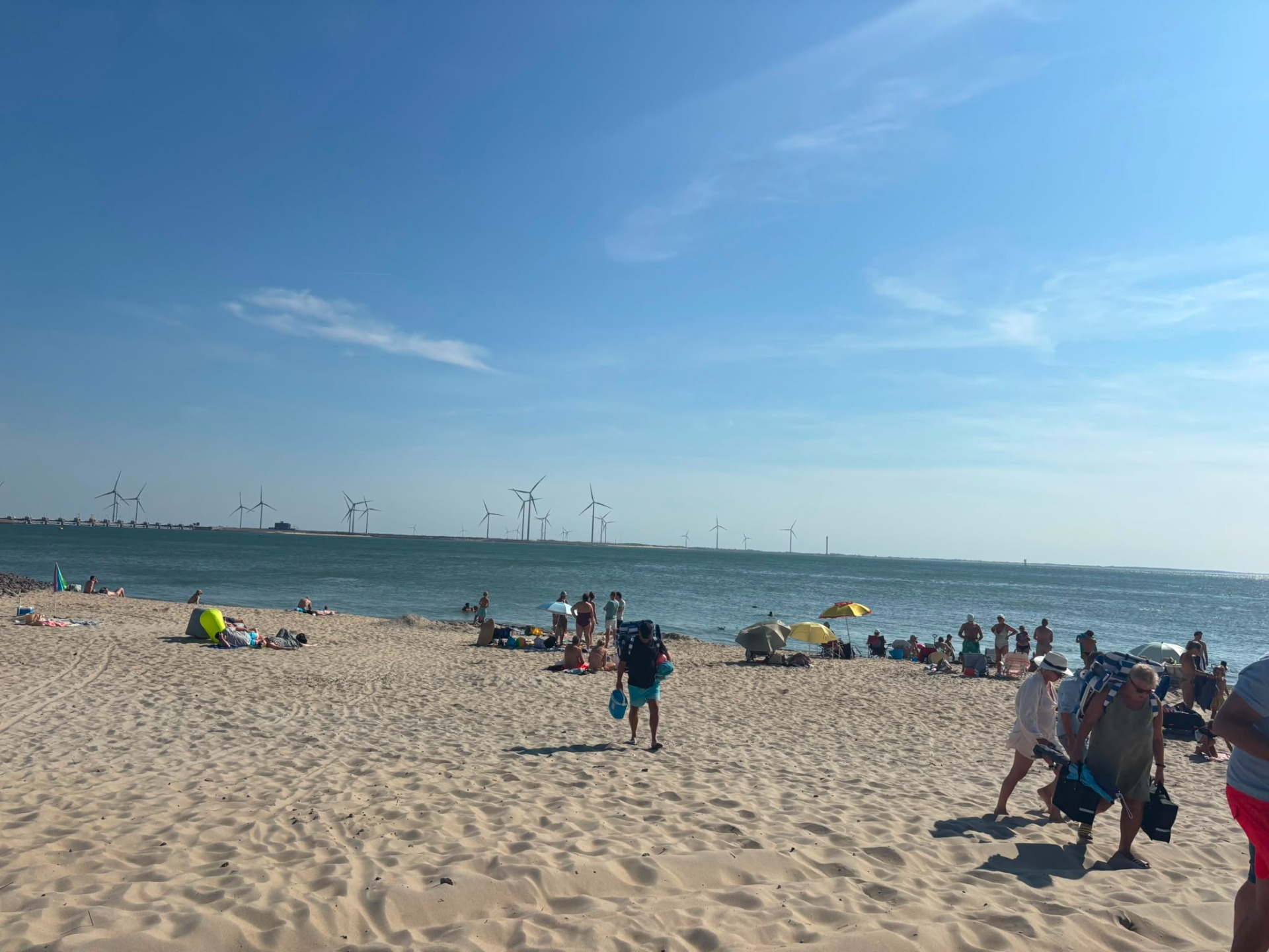 Zandstrand met badgasten, parasols en windturbines in de verte onder een blauwe lucht.
