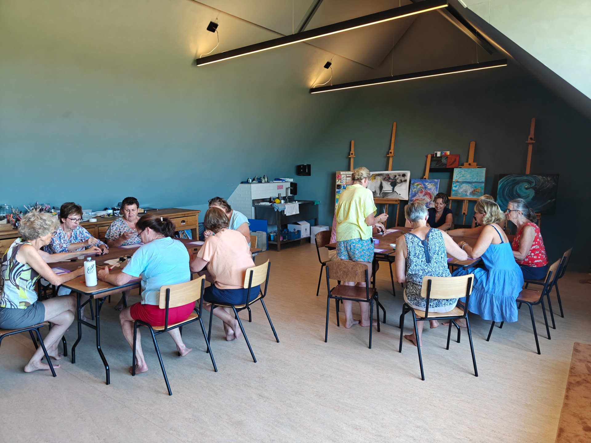 Kunstworkshop aan tafels, schilderijen en ezels, in een lichte ruimte.